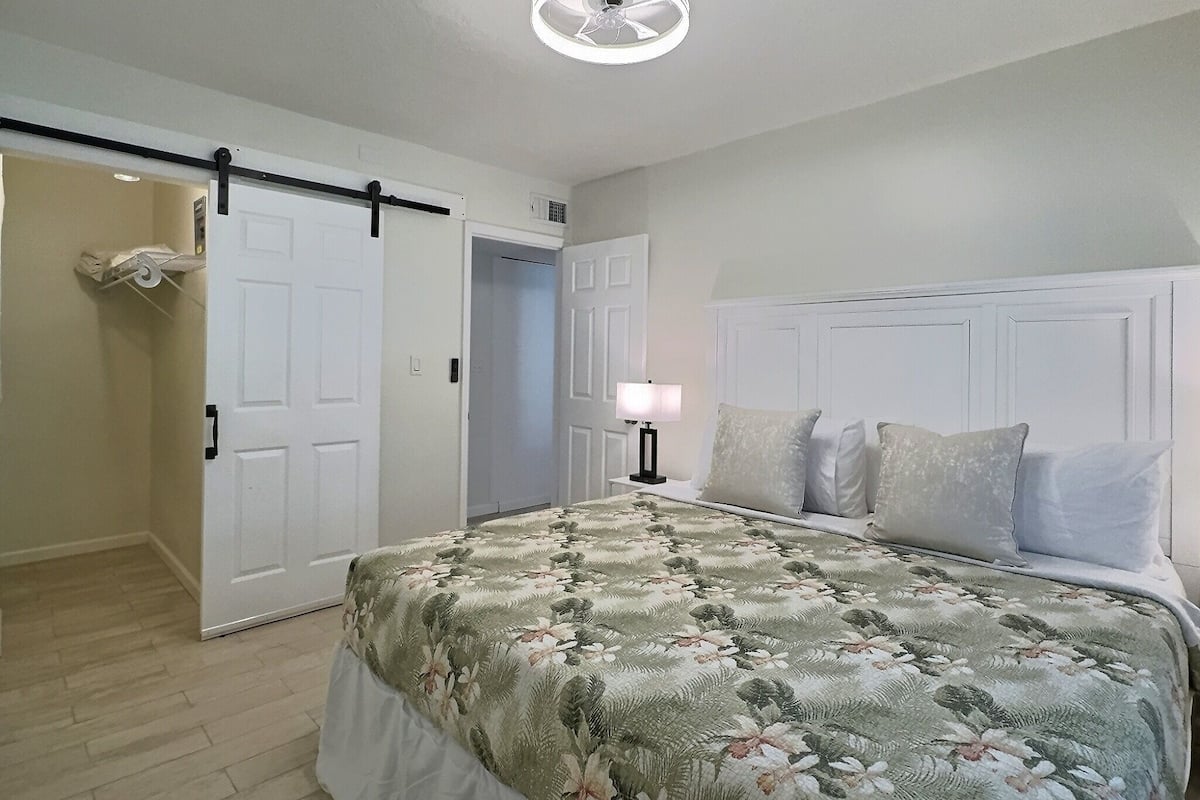 Une chambre à coucher comprenant un lit, une table de chevet, un ventilateur de plafond et une porte donnant sur une autre pièce.