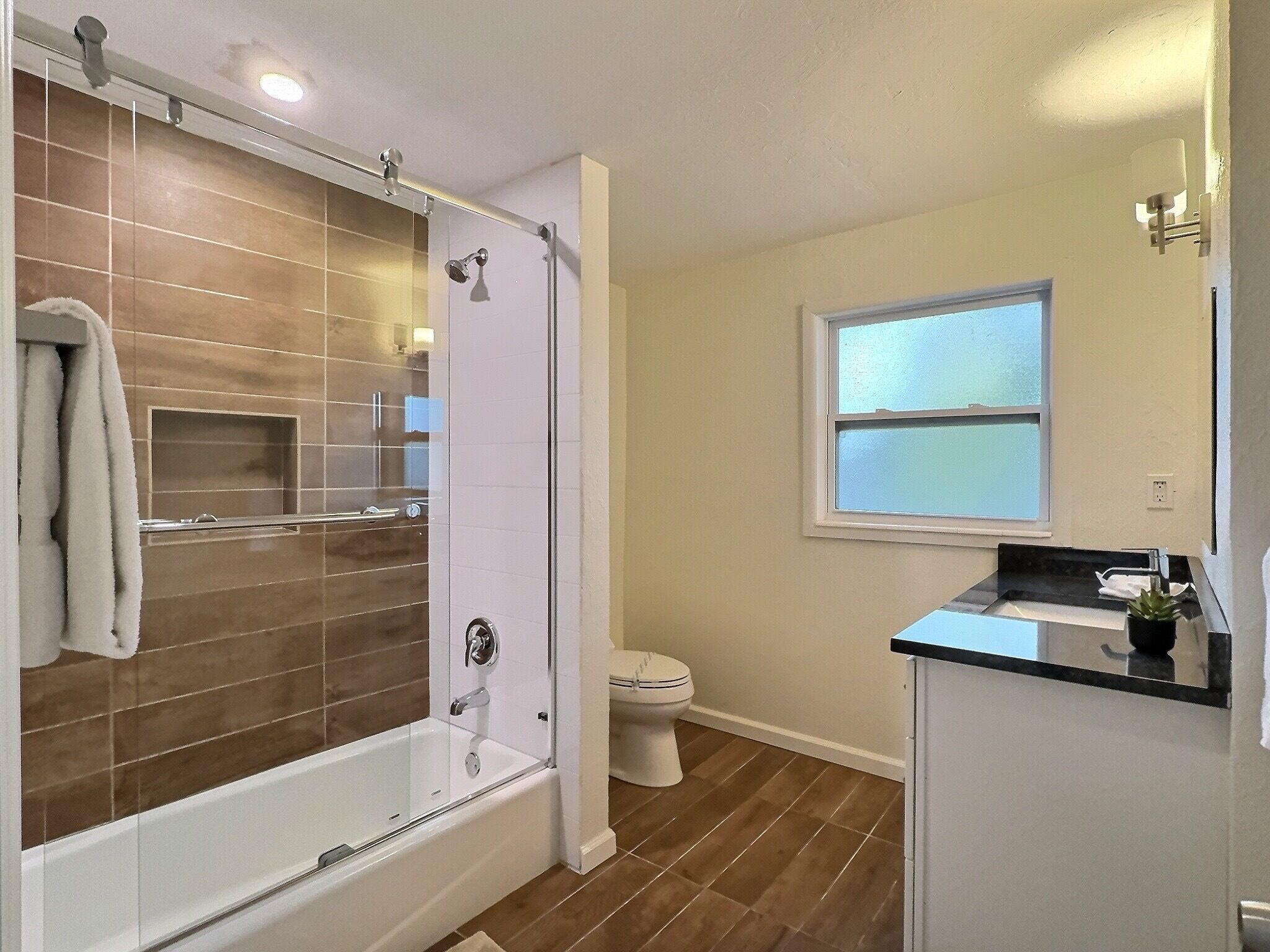 A modern bathroom with a glass-enclosed shower, a toilet, and a sink with a window above it.