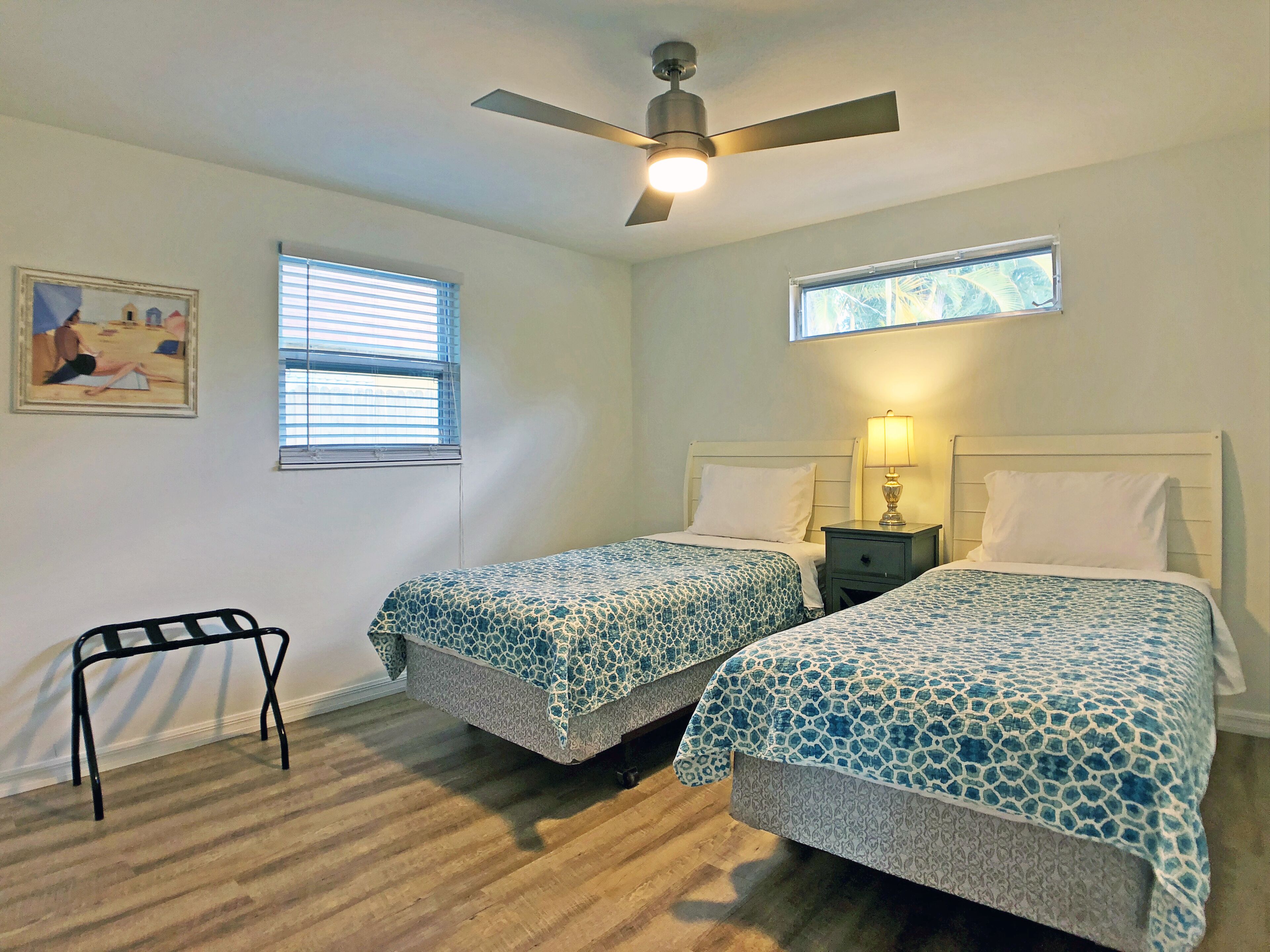 A bedroom with two beds, a ceiling fan, a nightstand with a lamp, a window with blinds, and a framed picture on the wall.