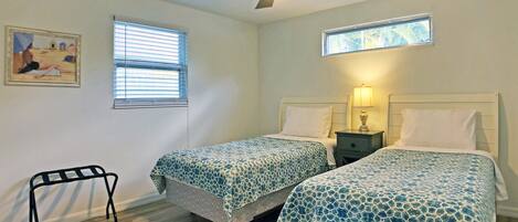 A bedroom with two beds, a ceiling fan, a nightstand with a lamp, a window with blinds, and a framed picture on the wall.