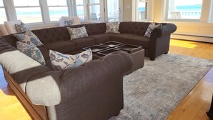 A living room with a large sectional sofa, a coffee table, and a view of the beach through large windows. - Luxurious Beachfront House - spectacular ocean views - w/ indoor elevator (Old Orchard Beach)
