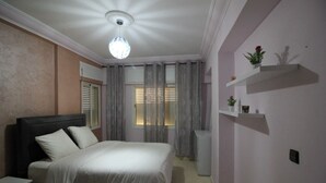 Quarto com cama, mesa de cabeceira, cômoda, luminária no teto, janela com cortinas e prateleira na parede com objetos decorativos.
