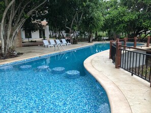 A swimming pool with a tiled edge and a curved design, surrounded by a concrete deck and a metal fence. - A comfortable and beautiful apartment (Puerto Morelos)