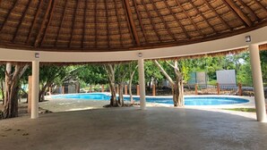 Ein überdachter Außenbereich mit einem reetgedeckten Dach, einem Betonboden und einem Pool im Hintergrund.