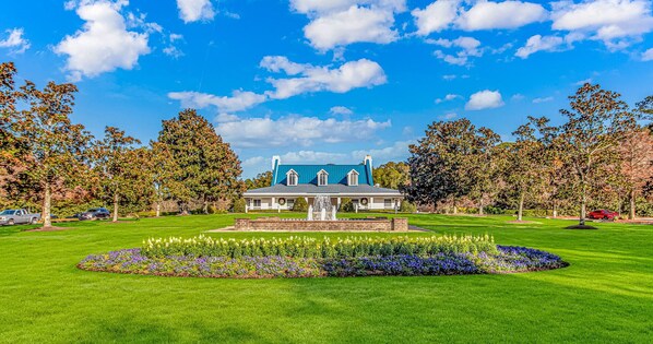 A large house with a fountain in the front lawn, surrounded by trees and a clear blue sky.