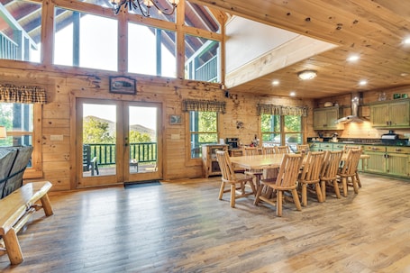 A wooden cabin interior with a dining area, kitchen, and large windows.