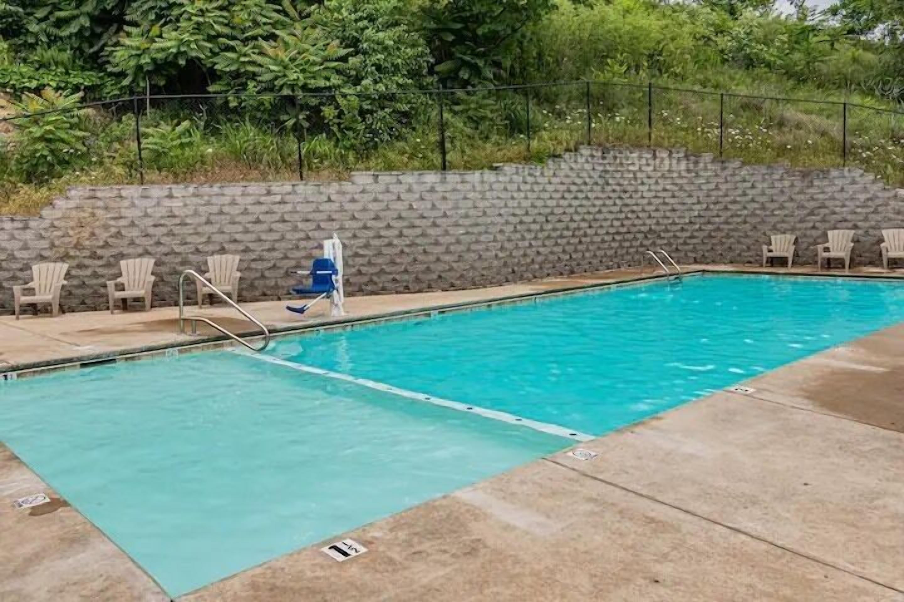 A rectangular swimming pool with clear blue water, surrounded by a concrete deck. There are several lounge chairs along the poolside, and a stone wall with greenery in the background.