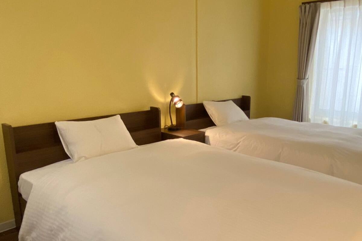 A hotel room with two single beds, a nightstand with a lamp, and a window with curtains.