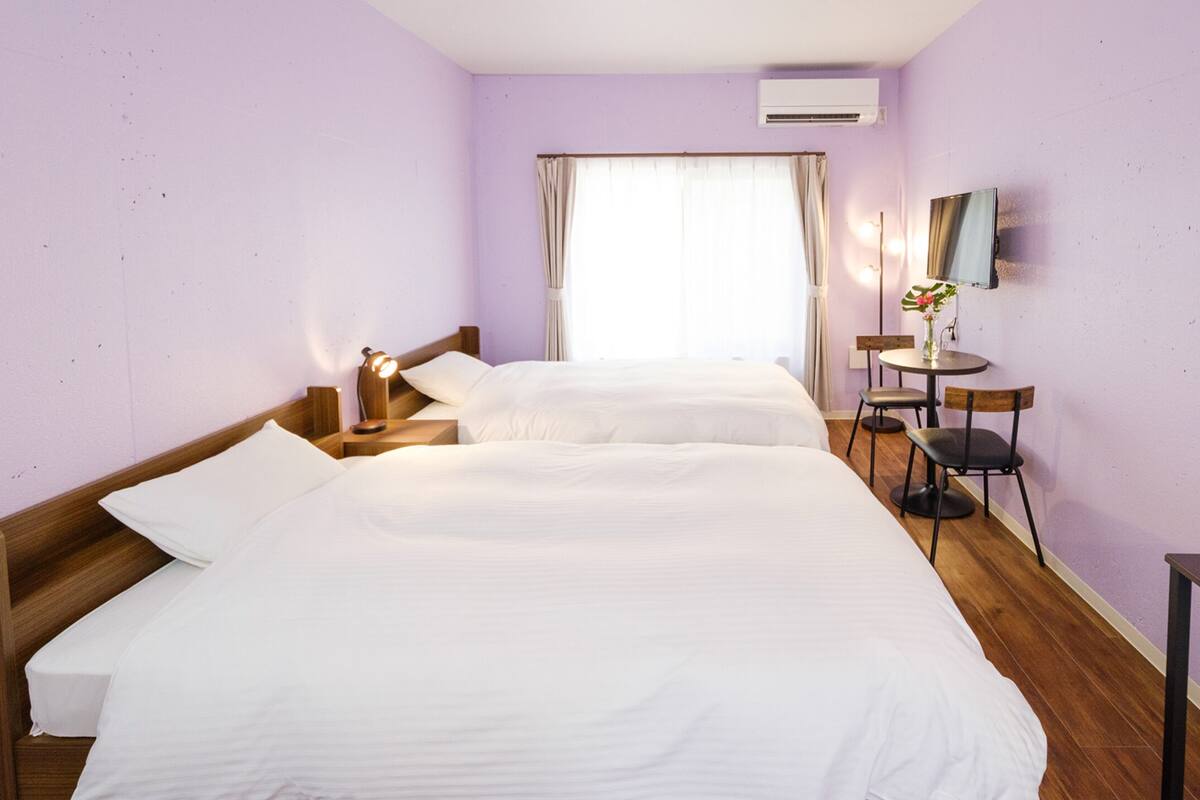 A hotel room with two single beds, a small round table with a chair, a TV mounted on the wall, and an air conditioning unit.