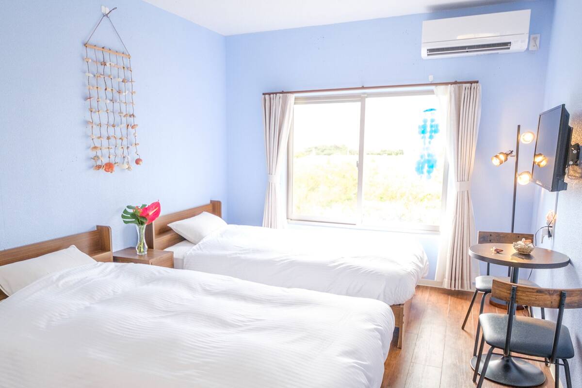 A hotel room with two beds, a small table with a chair, a decorative hanging, and a large window with curtains.