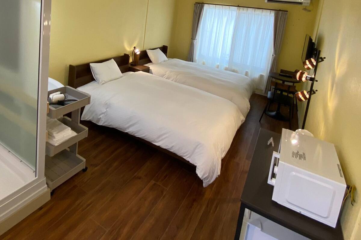 A hotel room with two beds, a small table with a lamp, a desk with a computer, and a shower area.