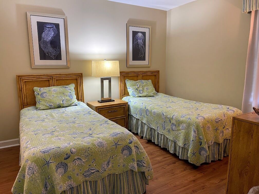A room with two single beds, a nightstand with a lamp, and framed artwork on the walls.