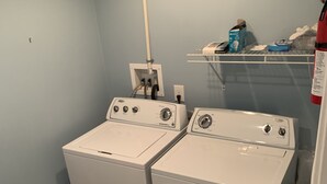 A laundry area with two white front-loading washers and dryers, a white pipe on the wall, and a shelf with various items. - Luxury beach house   (Seaside Heights)