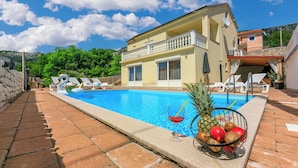 A poolside area with a pool, lounge chairs, and a fruit bowl with tropical fruits and a drink. - Villa Marta (Grizane)