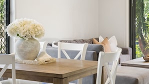Ein modernes Wohnzimmer mit einem Holzesstisch, weißen Stühlen, einem grauen Sofa mit Kissen und einer Vase mit weißen Blumen auf dem Tisch.