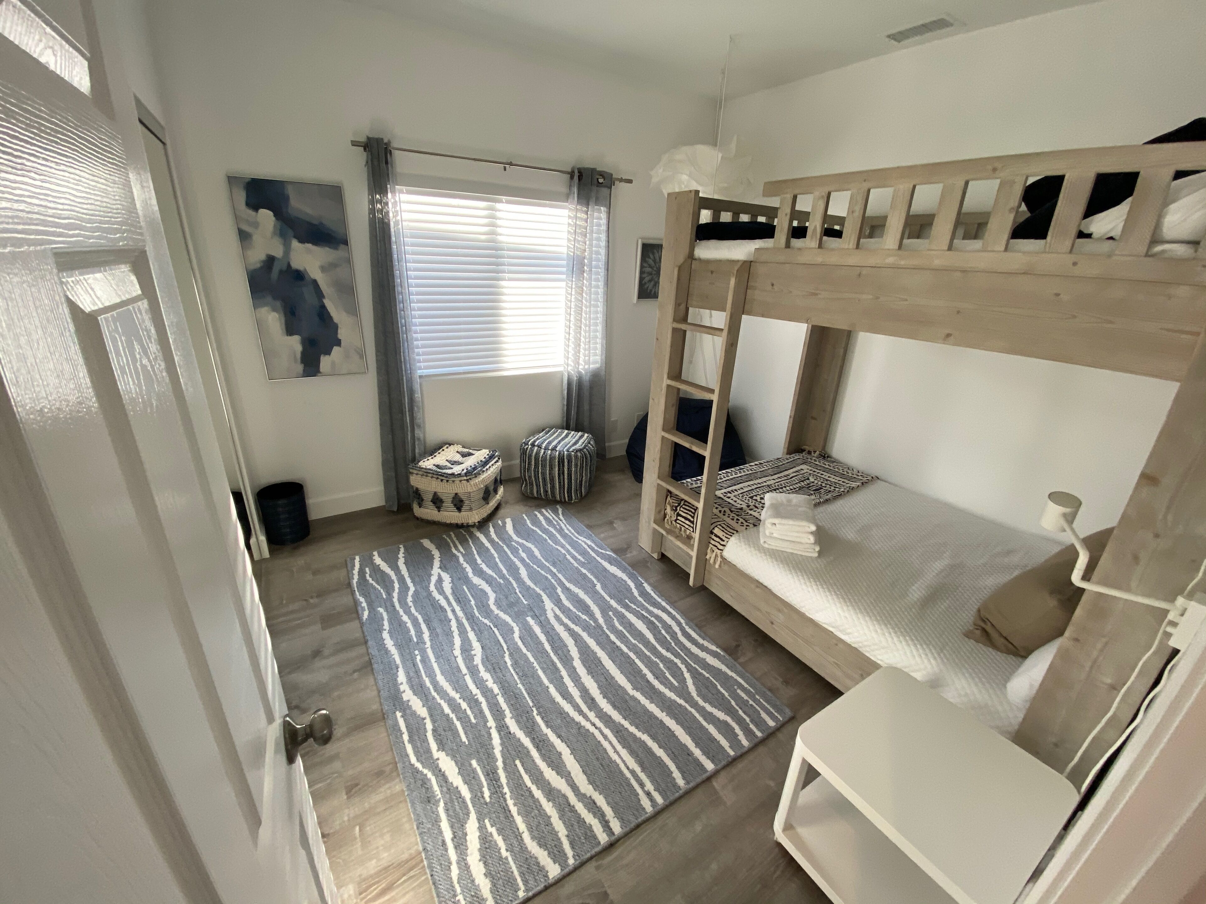 Une chambre à coucher de taille modeste, avec un lit superposé, une petite table blanche, un tapis à motifs et une fenêtre avec des rideaux.