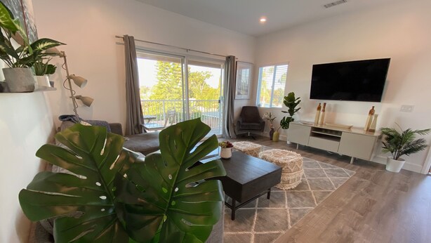 A modern living room with a dark wooden coffee table, a sofa, and a TV mounted on the wall. There are large windows with curtains, a balcony, and several potted plants. - *Cali King Bed * Bright Natural Light * Tall Ceilings * Full Kitchen * Balcony (San Diego)
