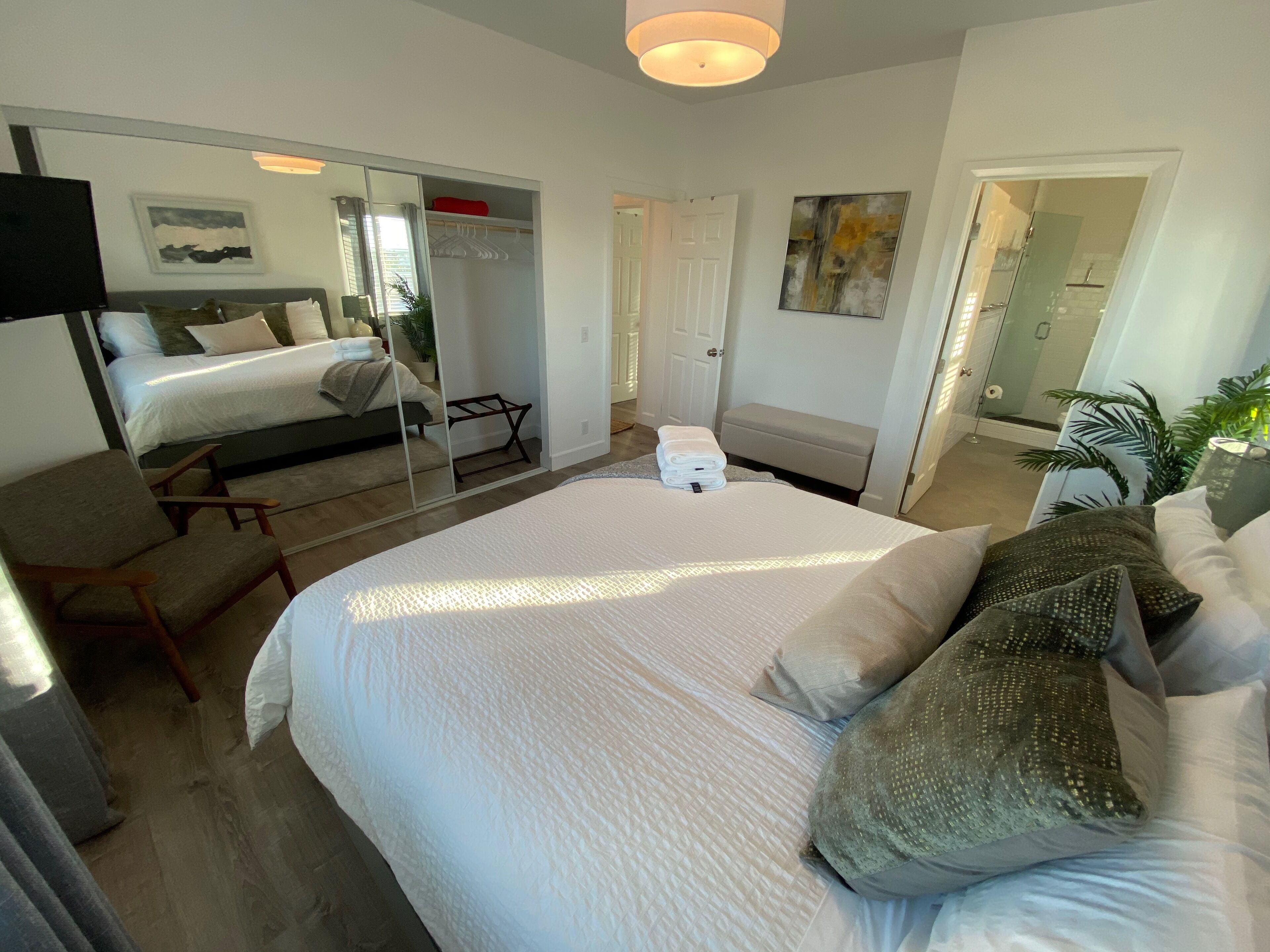 Une chambre d’hôtel moderne avec un grand lit, un coin salon comprenant une chaise, une petite table et un miroir. Une porte donne accès à une salle de bain et à un espace douche.