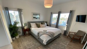 A modern bedroom with a large bed, a bedside table with a lamp, a chair, a TV mounted on the wall, and a window with curtains. - *Cali King Bed * Bright Natural Light * Tall Ceilings * Full Kitchen * Balcony (San Diego)