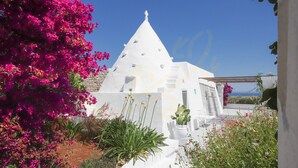 Ein weißes Gebäude mit einem spitzen Kegeldach und einem Kreuz darauf, umgeben von rosa Bougainvillea und Grünpflanzen.