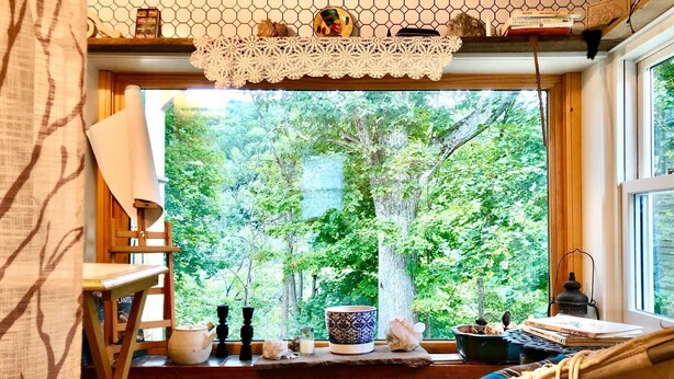 Una habitación acogedora con un amplio ventanal que da a un exuberante jardín, un banco de madera y una mesa con diversos objetos.