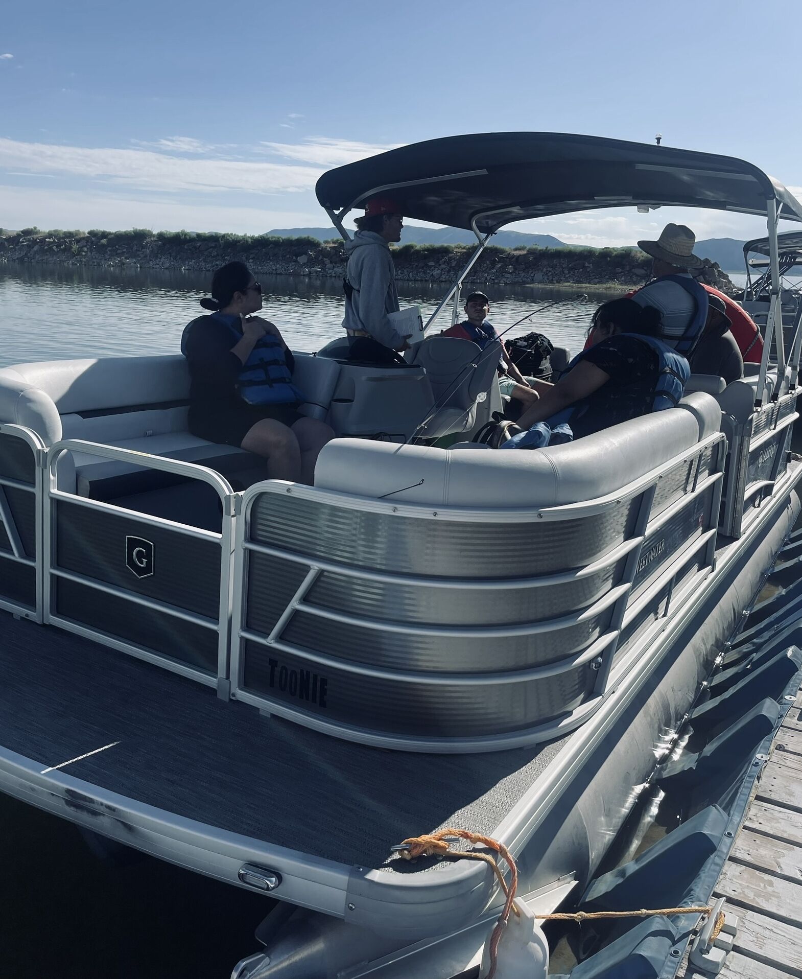 A pontoon boat docked at a pier with passengers seated and a person standing at the helm.