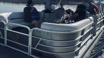 A pontoon boat docked at a pier with passengers seated and a person standing at the helm.