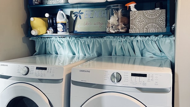 Un espace buanderie équipé de deux machines à laver Samsung, d’une étagère avec des serviettes et d’un panneau indiquant : « LA PLAGE, c’est par là ».
