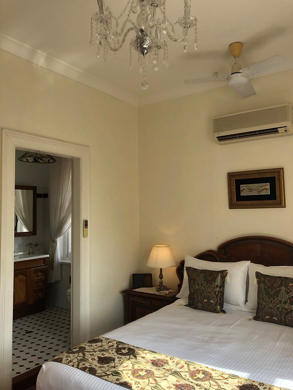 A hotel room with a bed, a chandelier, an air conditioner, a bedside lamp, and a framed picture on the wall.