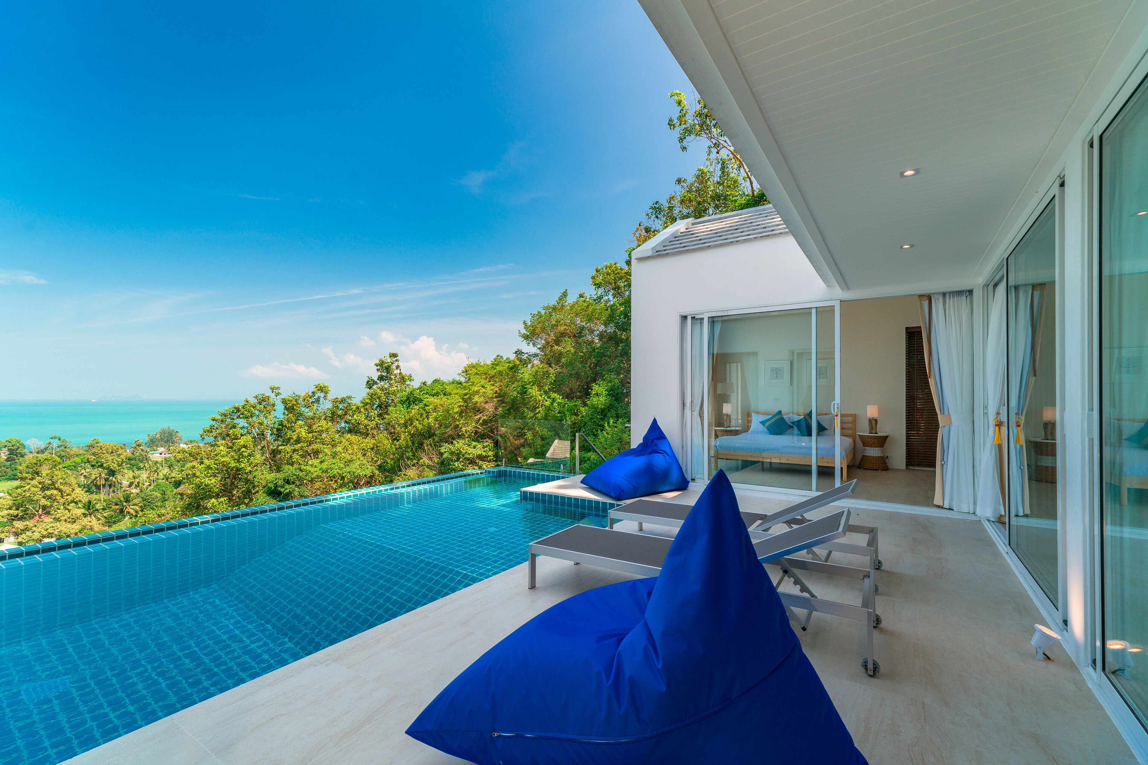 Un'area piscina moderna con vista aperta sull'oceano, una stanza vetrata con un letto e una poltrona da relax con cuscino blu.