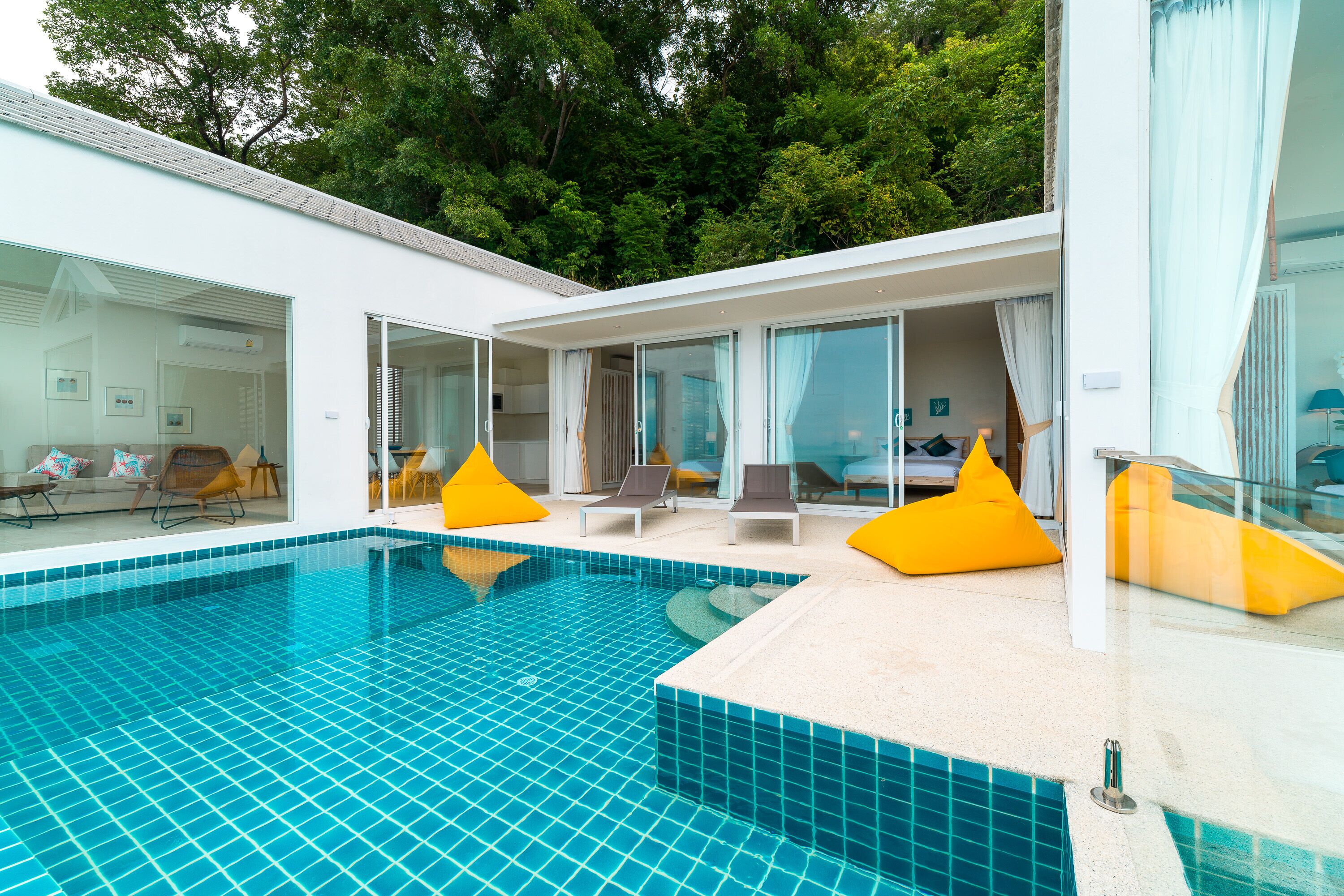 A modern pool area with clear blue tiles, yellow bean bag chairs, and a glass-enclosed room with a sofa and chairs.