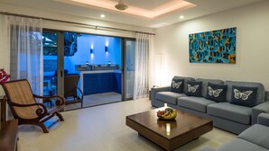 A modern living room with a sofa, a wooden coffee table with fruit, a wicker chair, and a view of a kitchen and balcony through glass doors. - Modern Tropical Villa (Koh Samui)
