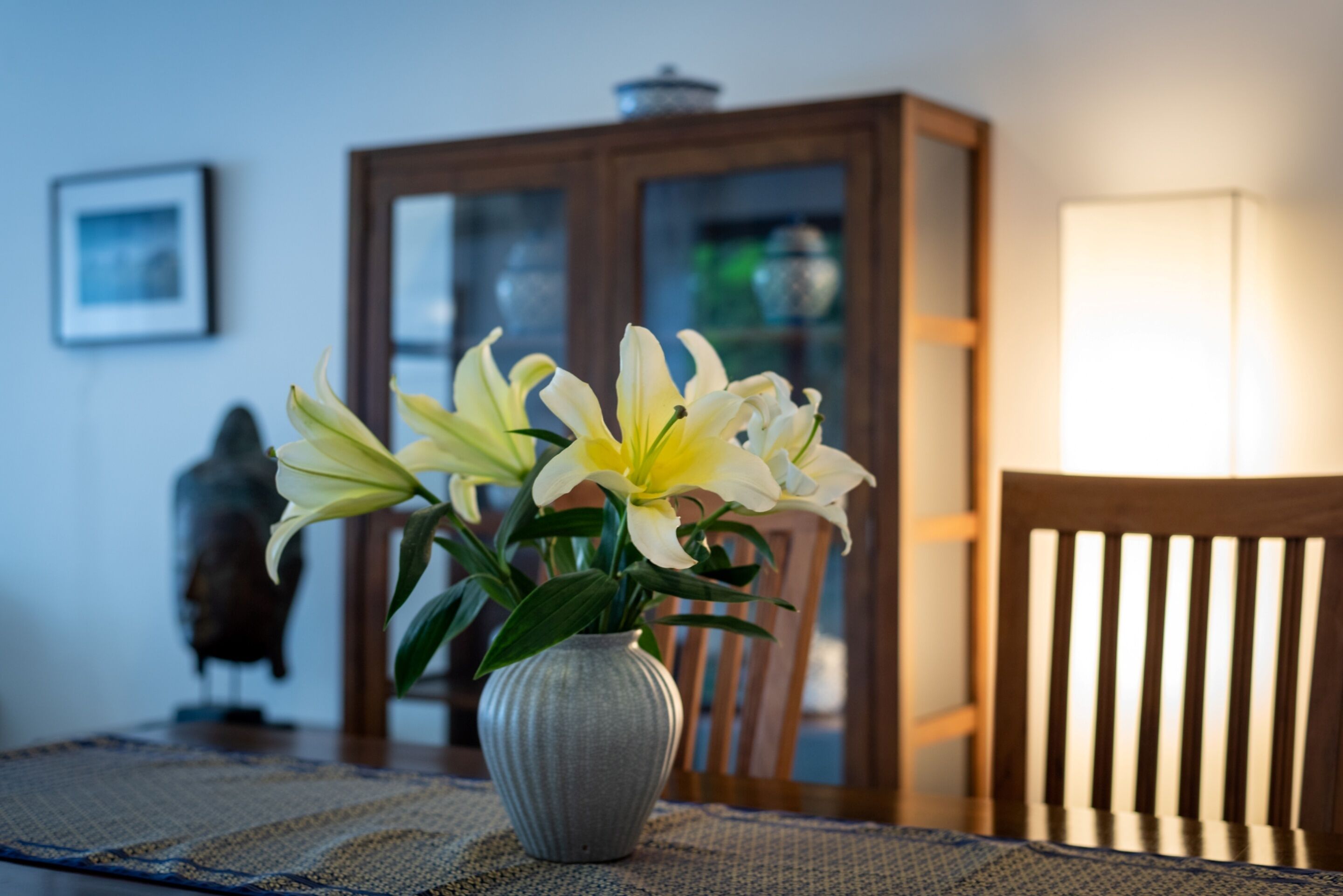 A dining table with a vase of yellow lilies, a wooden chair, and a cabinet in the background.