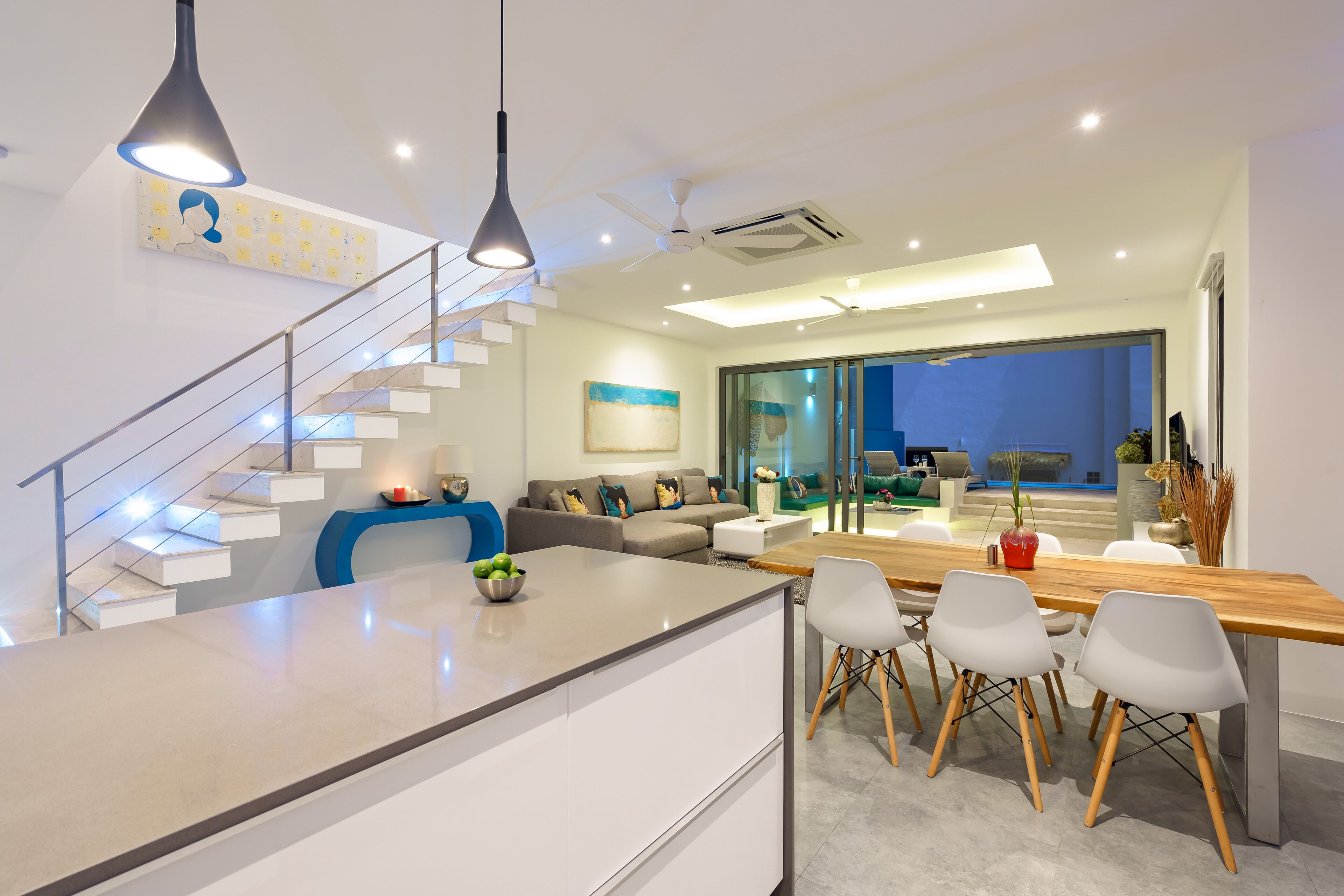 Une cuisine moderne dotée d’un îlot central, un coin repas avec une table et des chaises en bois, un escalier avec une rampe en verre et un salon avec un canapé et une table basse.