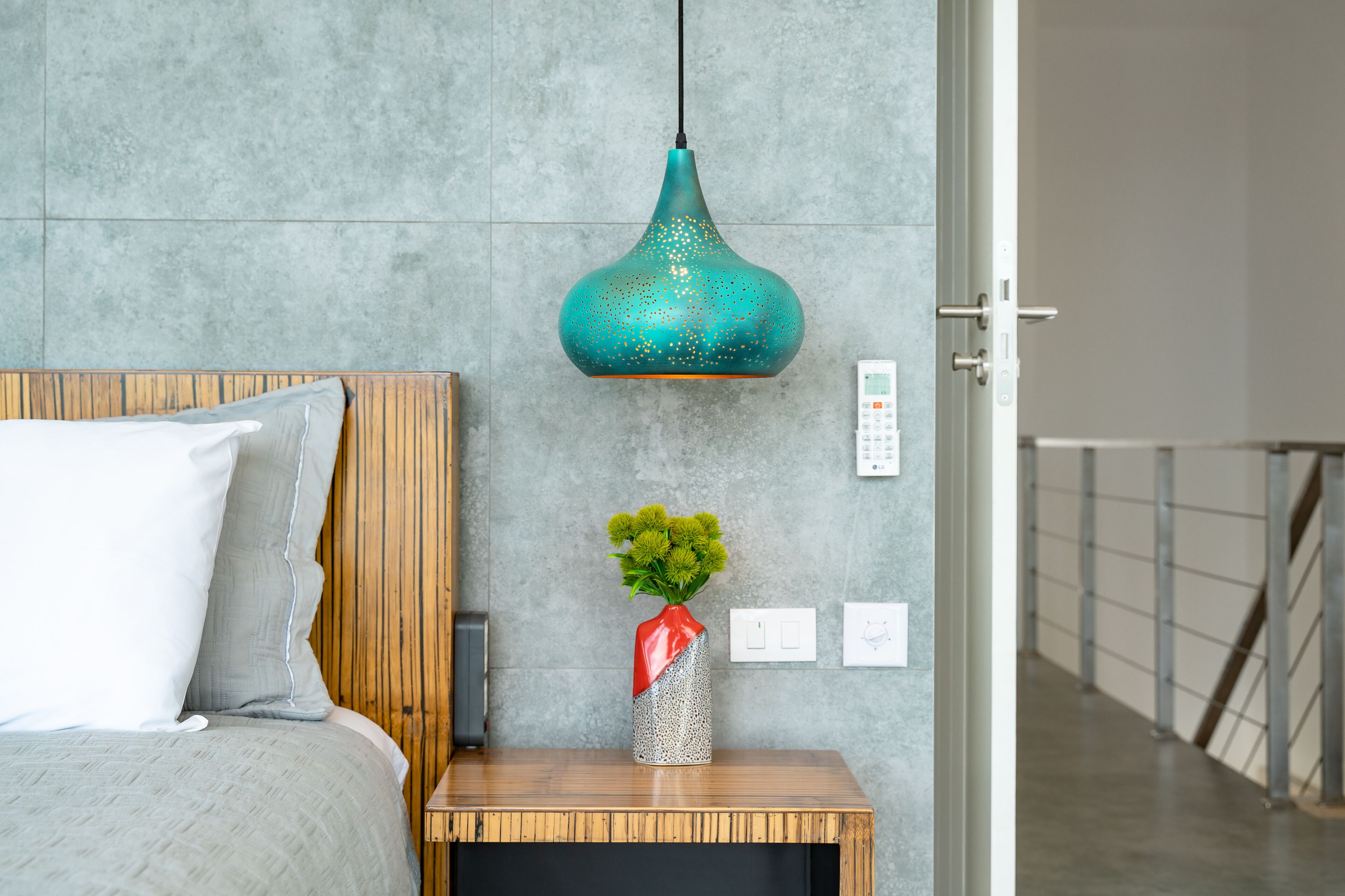 A bedroom with a bed, a nightstand with a vase of flowers, a teal pendant light, and a door to another room.