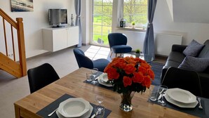 A dining area with a wooden table set for four, a vase of orange flowers, and a television on a white cabinet. - 3-room maisonette apartment 13 (AF) - Lancken-Granitz apartment complex (Lancken-Granitz)