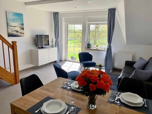 A dining area with a wooden table set for four, a vase of orange flowers, and a television on a white cabinet. - 3-room maisonette 14 (AF) - Apartment complex Lancken-Granitz (Lancken-Granitz)
