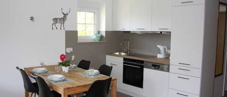 Une cuisine moderne avec des meubles blancs, une table à manger en bois, des chaises noires et une petite fenêtre donnant sur un espace verdoyant.