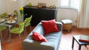 Een woonkamer met een grijze bank, rode kussens, een eettafel met stoelen, een zwarte kast en een raam met gordijnen.