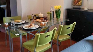 Een eetgedeelte met een glazen tafel, groene stoelen, een bank, een lamp en een decoratief wandobject.