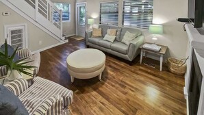 A spacious living area with a wooden floor, a grey sofa, a round ottoman, and a staircase.