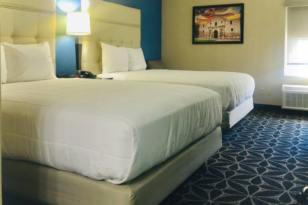 A hotel room with two beds, a nightstand, a lamp, a painting, and a window.