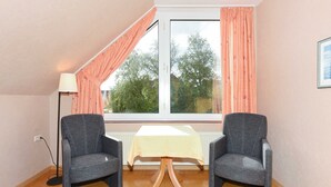 Une pièce avec deux fauteuils gris, une petite table recouverte d’une nappe jaune, un lampadaire et une fenêtre ornée de rideaux roses.