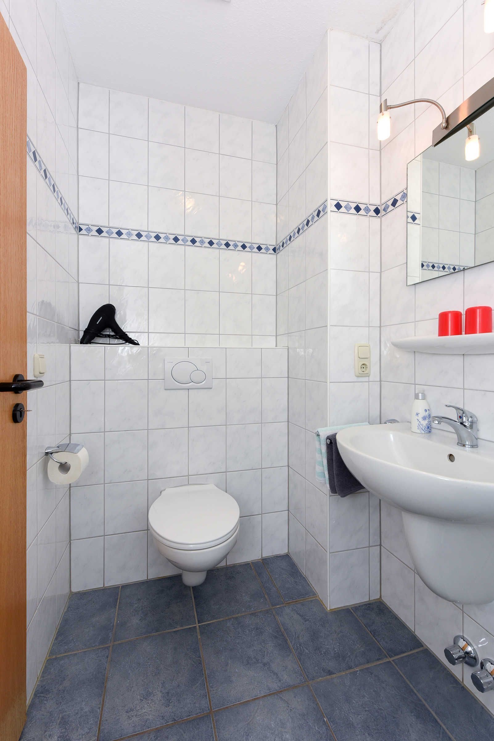 A bathroom with white tiles, a white toilet, a sink with red toiletries, and a towel hanging on the wall.
