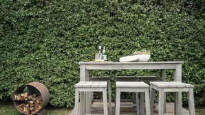 A wooden outdoor table with two stools, a planter with plants, and a log storage barrel.
