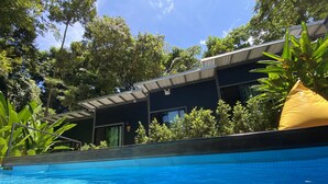 Una piscina con agua azul cristalina, rodeada de abundante vegetación y un edificio moderno con grandes ventanales y techo plano.