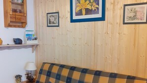 A cozy living room with a plaid sofa, a small table with a lamp, and framed floral artwork on the wall. - Charming Studio in Verdant Setting with Balcony, Sleeps 4 (Le Biot)