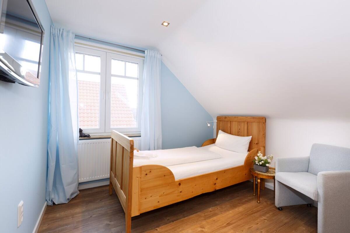 Een knus, klein slaapkamer met een houten bed, een raam met gordijnen, een radiator en een klein tafeltje met een bloevas.