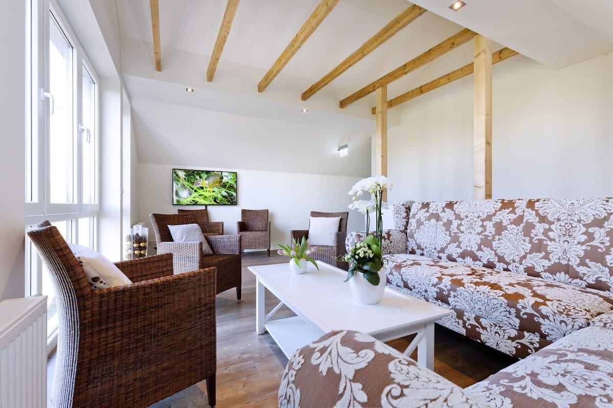 Een ruime woonkamer met rieten meubels, een witte bank met bloemenprint en een aan de muur bevestigde televisie.