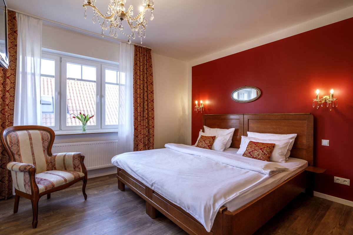 Een slaapkamer met een groot bed, een houten hoofdbord, een stoel, een raam met gordijnen en een kroonluchter.